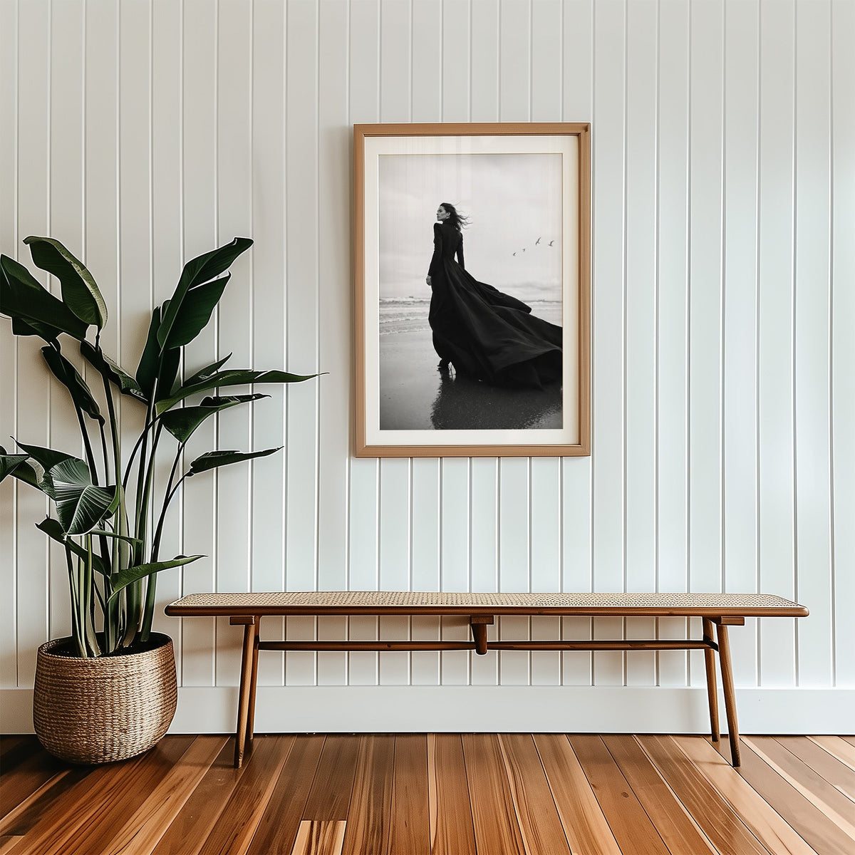 Woman in long black gown on a windy beach print in a wood frame with a white mat above a cane bench