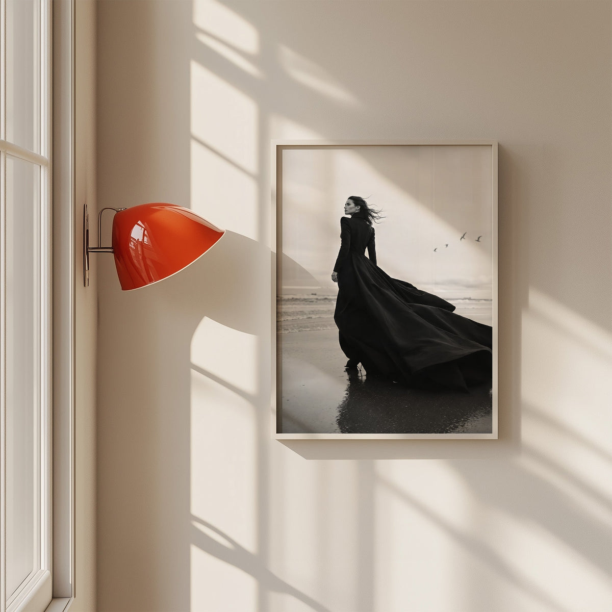 Woman in long black gown on a windy beach print in a white frame on a sunlit wall beside a red sconce