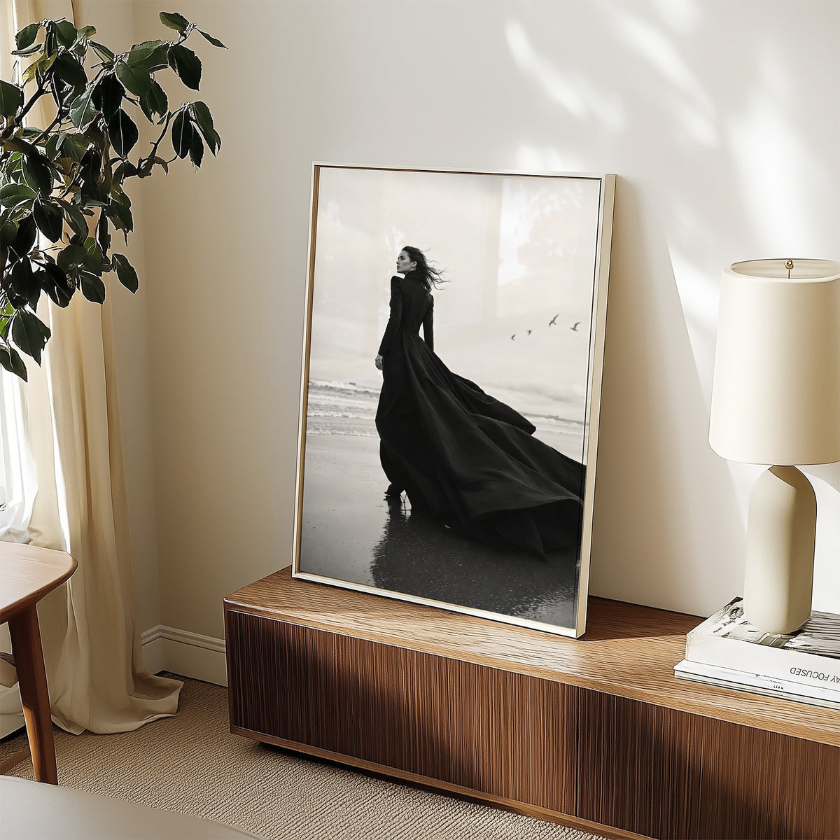 Woman in long black gown on a windy beach print in a white frame on a walnut sideboard beside a ceramic lamp