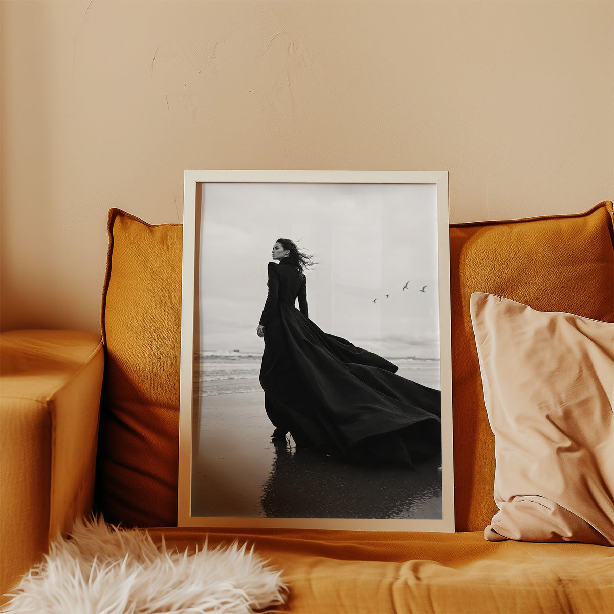 Woman in long black gown on a windy beach print in a white frame leaning on a mustard velvet sofa