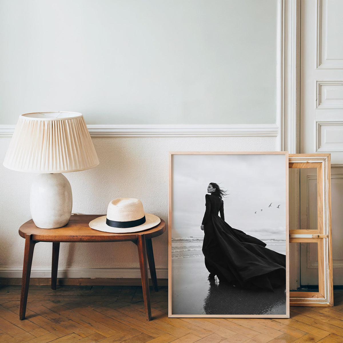 Woman in long black gown on a windy beach print in a pine frame on a wood floor beside a lamp and straw hat