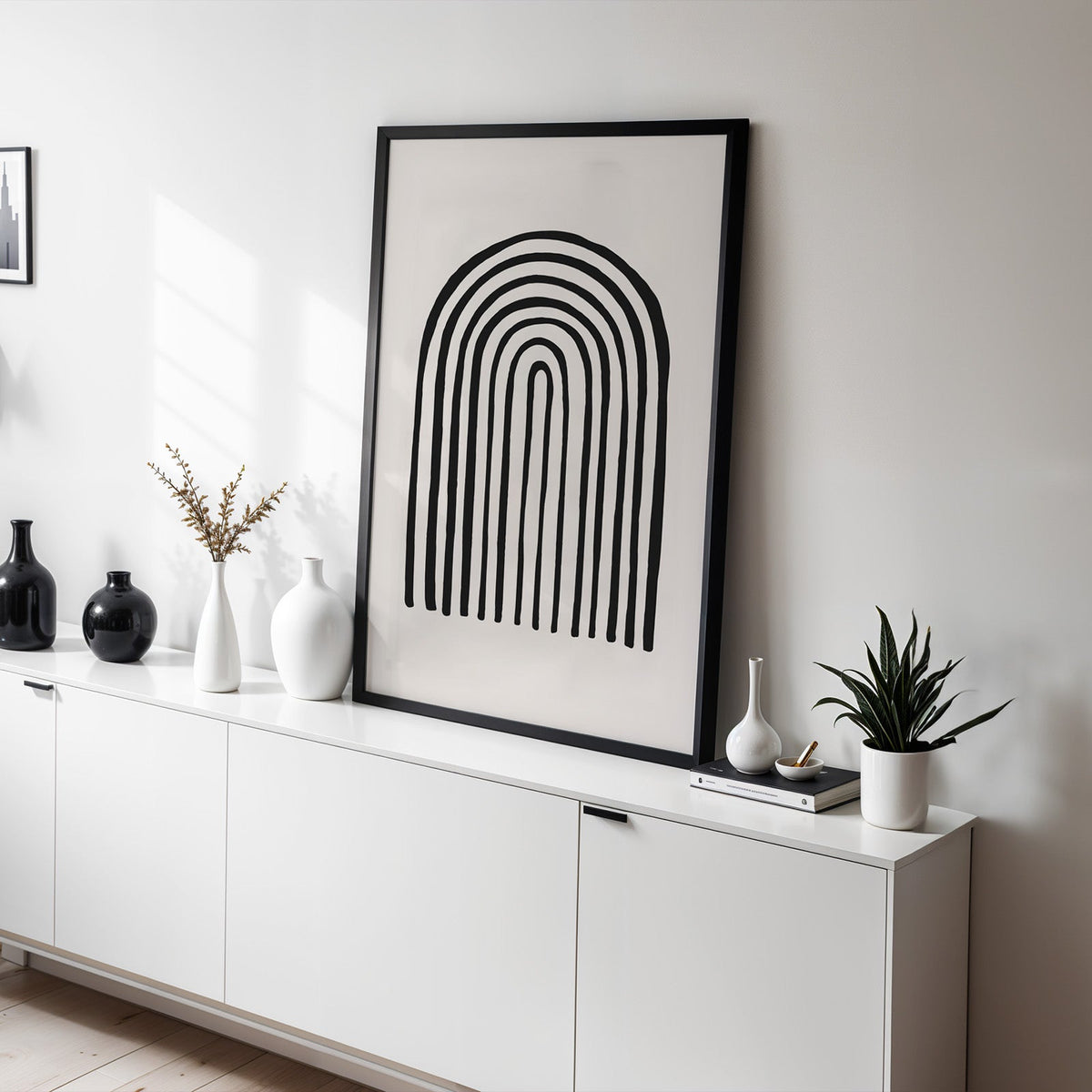 Black hand drawn rainbow arch line art print in a black frame leaning on a white sideboard with black and white vases