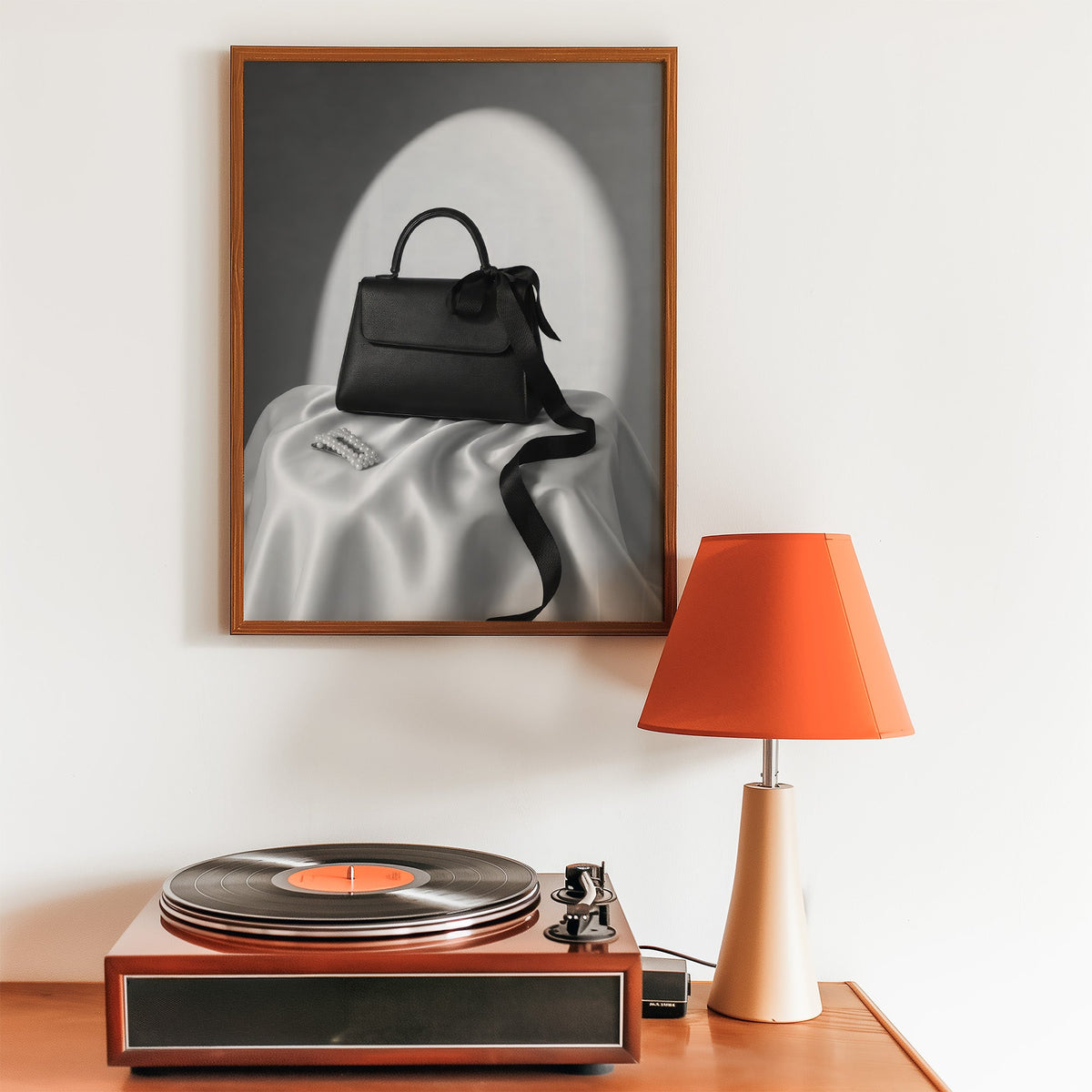 Black top-handle handbag on silk with pearl clip print in a walnut frame above a record player and orange lamp