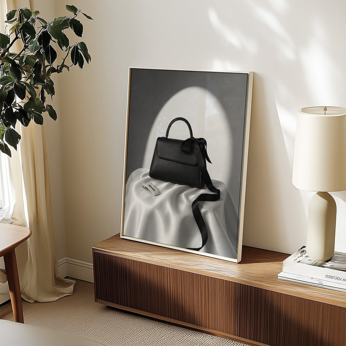 Black top-handle handbag on silk with pearl clip print in a white frame on a walnut sideboard beside a ceramic lamp