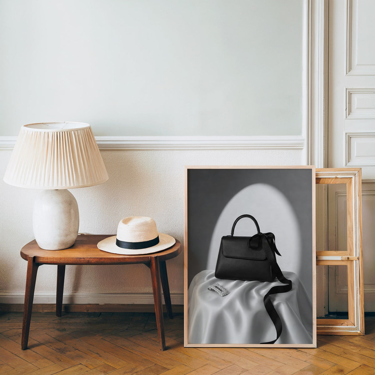 Black top-handle handbag on silk with pearl clip print in a pine frame on a wood floor beside a lamp and straw hat