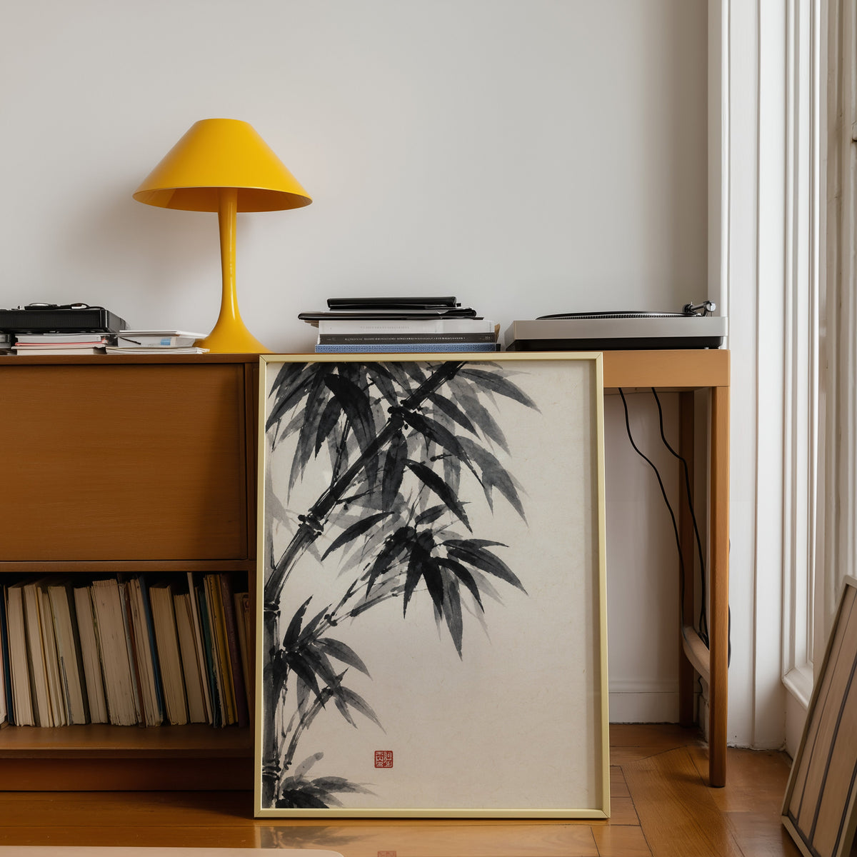 Black ink bamboo sumi-e poster shown in a gold frame leaning under a teak desk with a yellow lamp