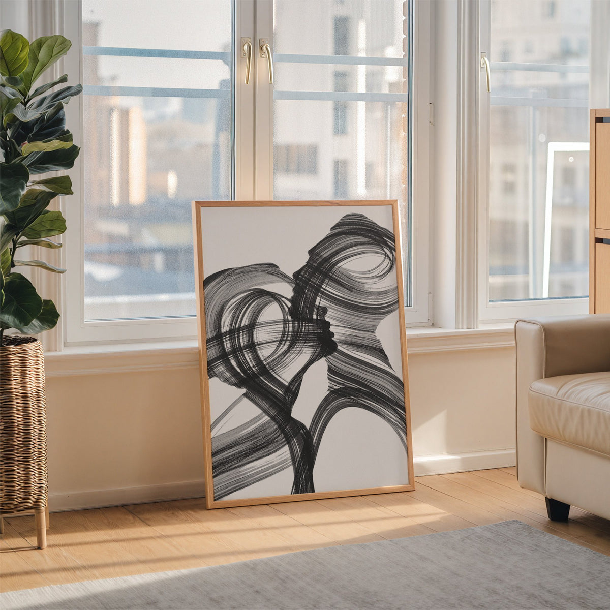 Black ink brush swirl abstract print in an oak frame leaning on the floor beside a fiddle leaf fig