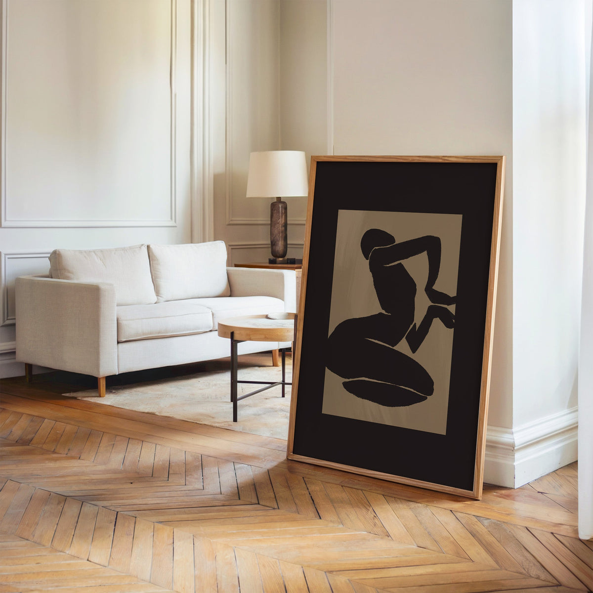 Black kneeling silhouette on taupe panel print in an oak frame leaning on a herringbone floor by a cream linen sofa
