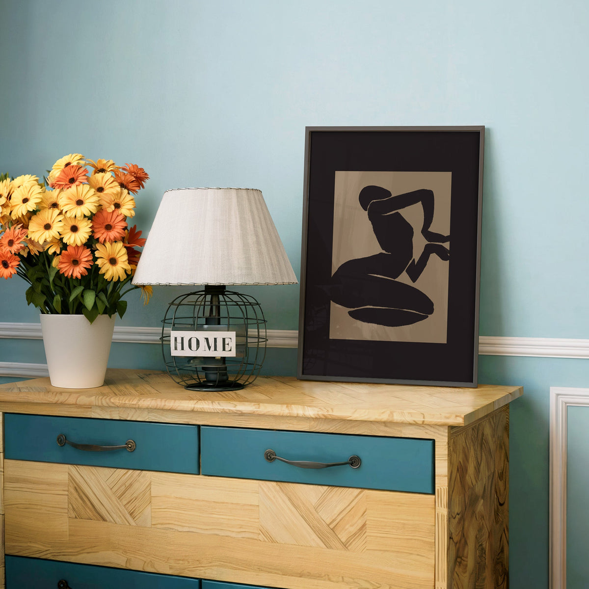 Black kneeling silhouette on taupe panel print in a charcoal frame on a teal dresser beside orange and yellow daisies