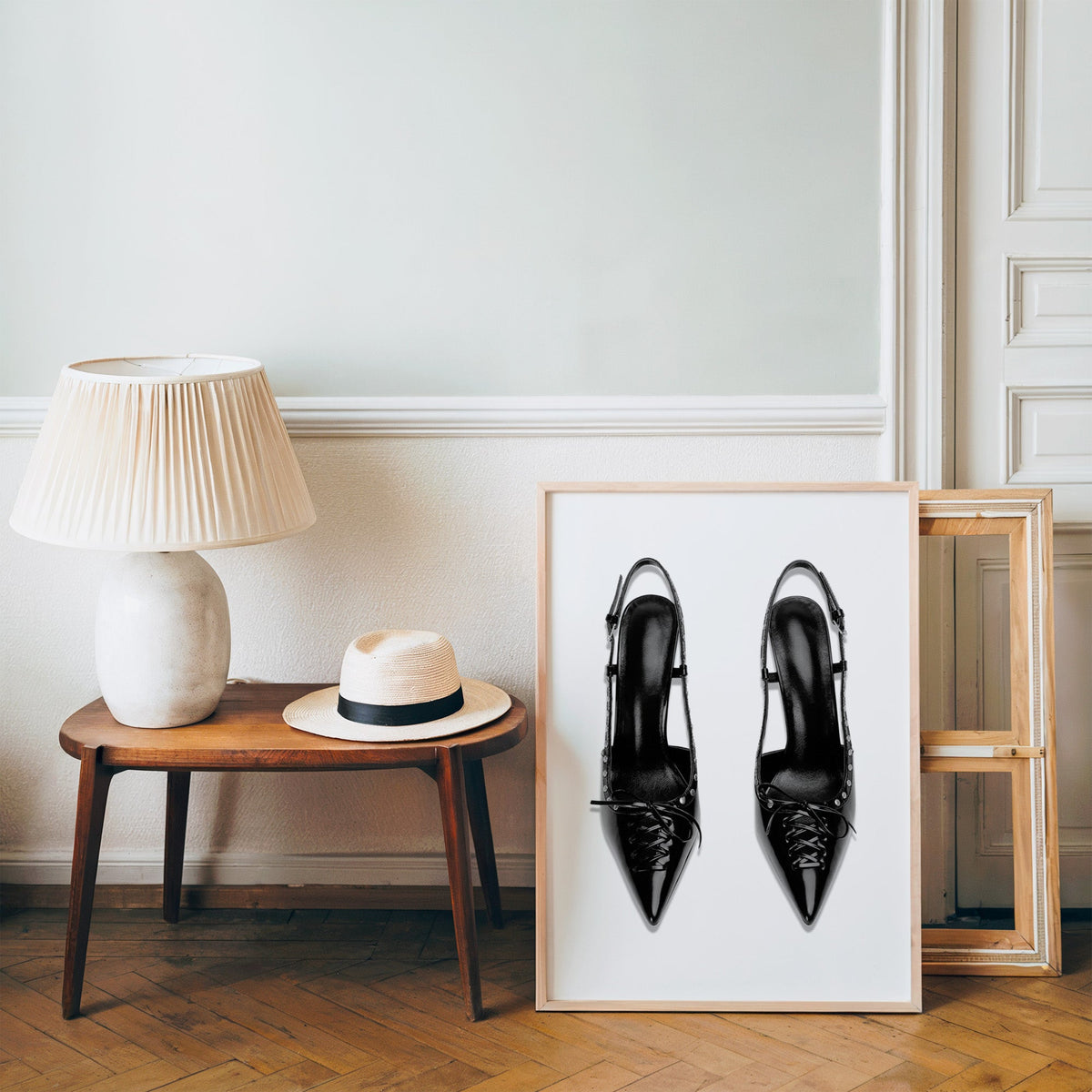 Black lace-up studded slingback heels print in a pine frame on a wood floor beside a lamp and straw hat