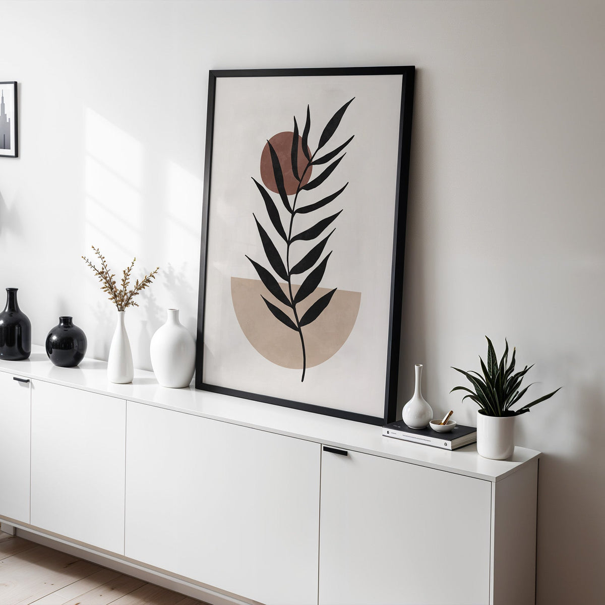 Black leaf branch with rust sun boho print in a black frame leaning on a white sideboard with black and white vases