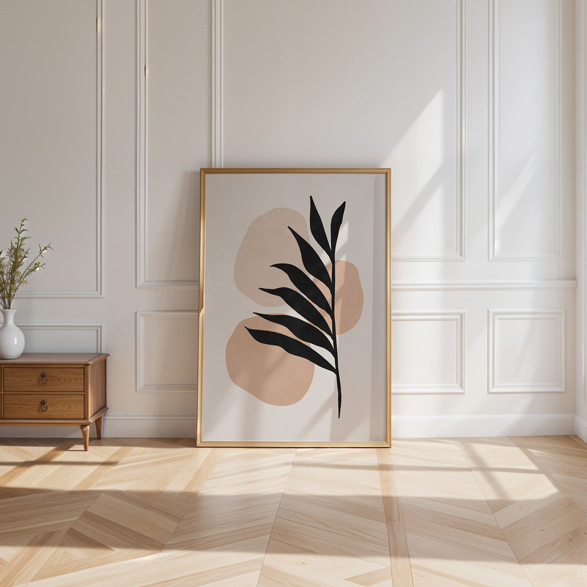 Black leaf over peach blobs boho print shown in a gold frame leaning on a paneled white wall in sunlight