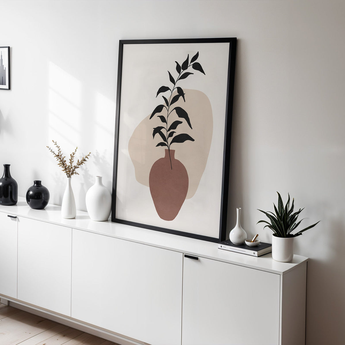 Black leaves in rosewood vase boho print in a black frame leaning on a white sideboard with black and white vases