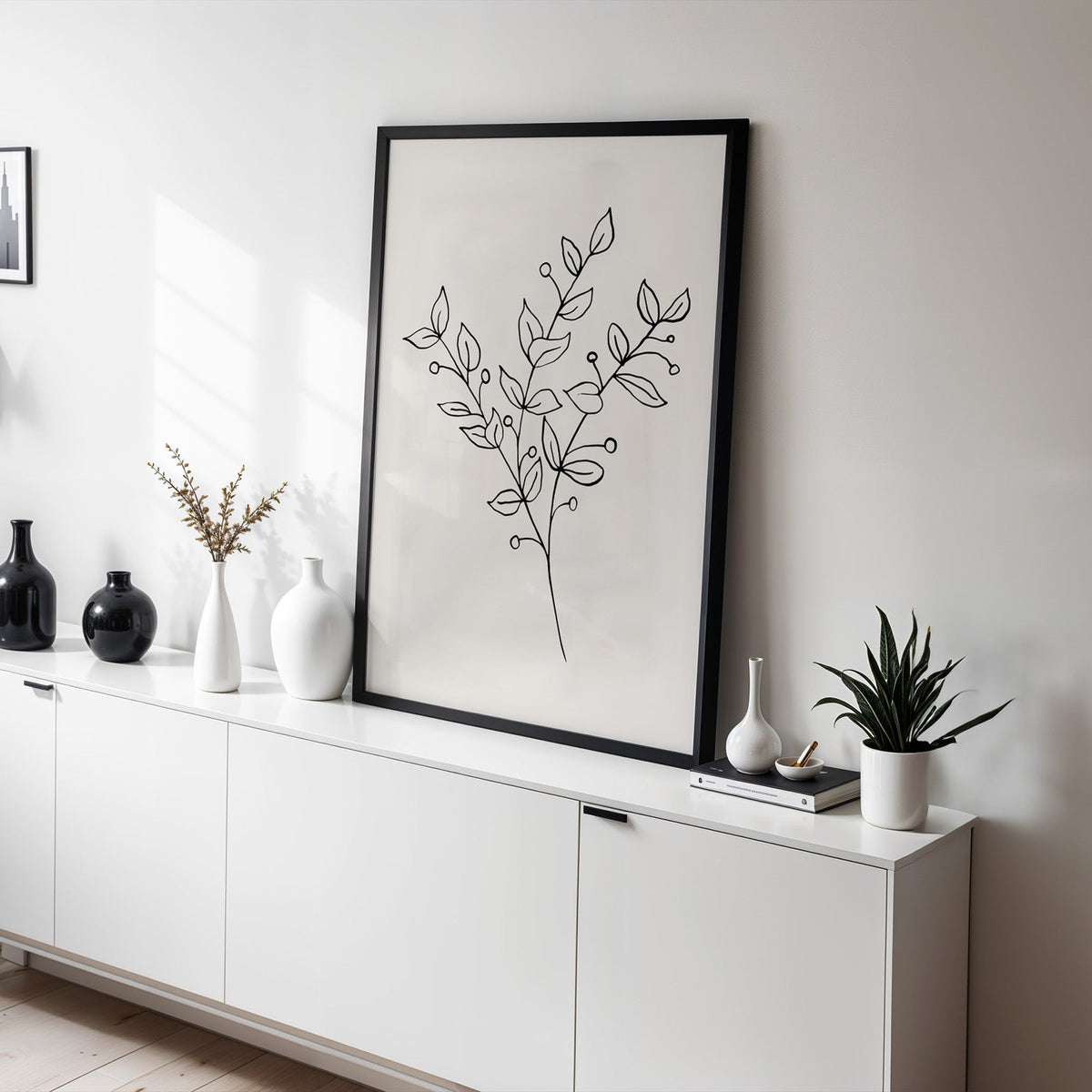 Black line berry sprig minimalist print in a black frame leaning on a white sideboard with black and white vases