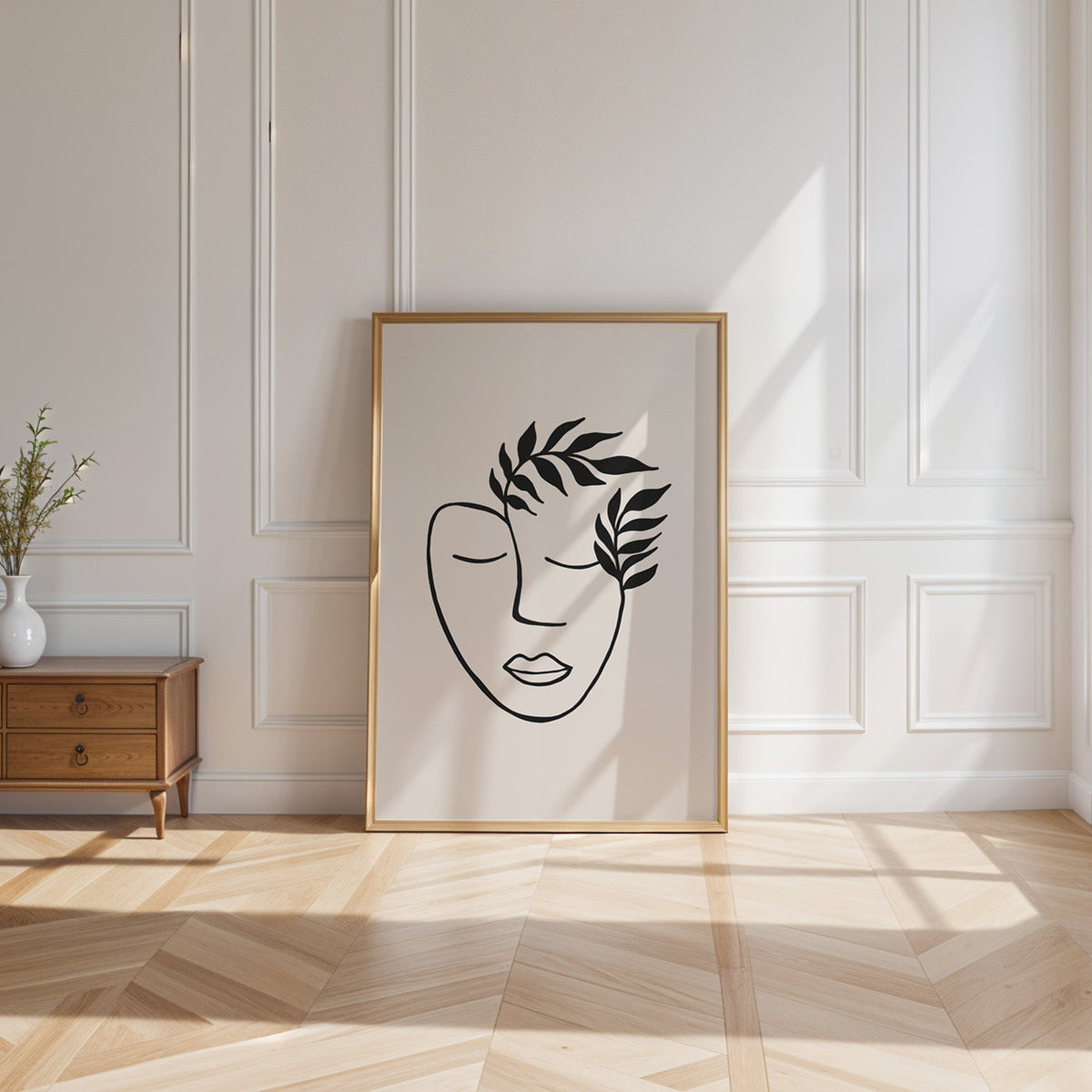 Black line face with leaf crown minimalist print shown in a gold frame leaning on a paneled white wall in sunlight