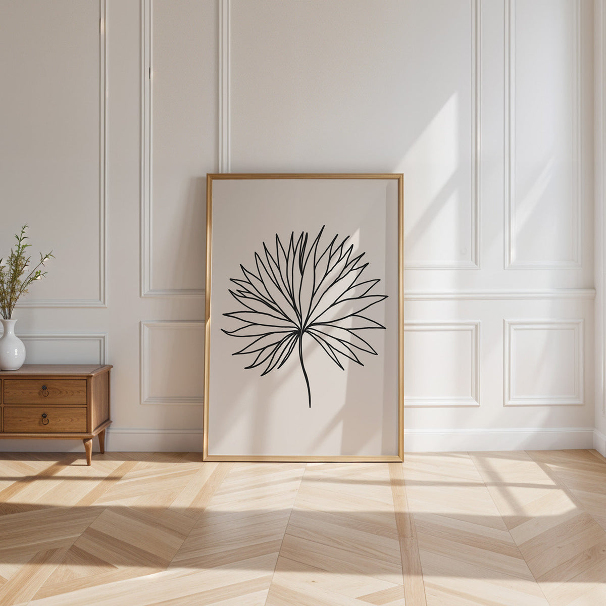 Black line fan leaf minimalist print shown in a gold frame leaning on a paneled white wall in sunlight