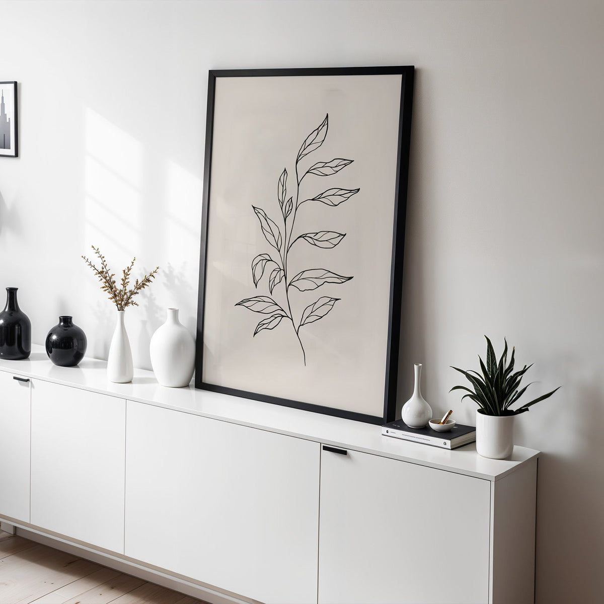 Black line leafy branch botanical line art print in a black frame leaning on a white sideboard with black and white vases