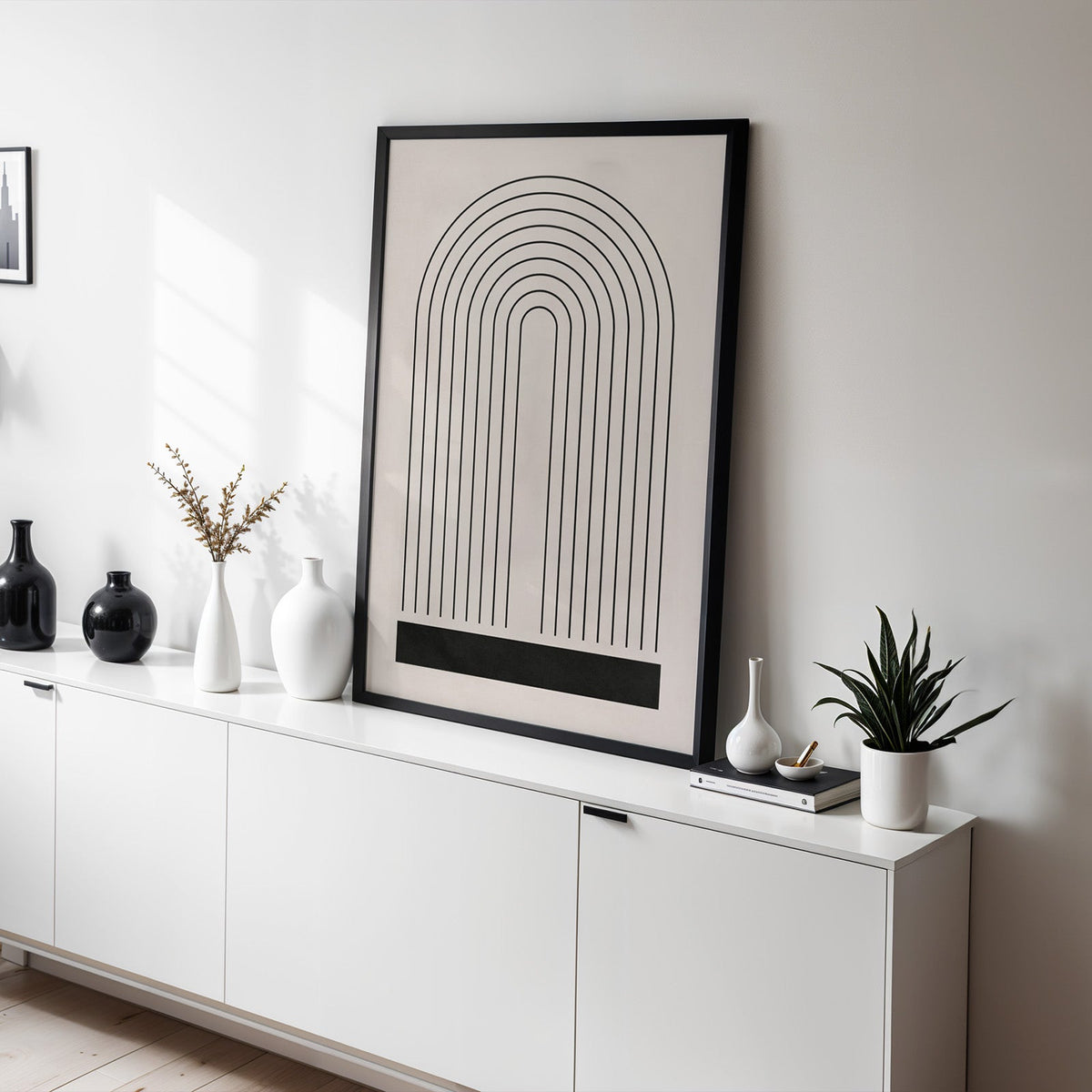 Black line rainbow arch minimalist print in a black frame leaning on a white sideboard with black and white vases