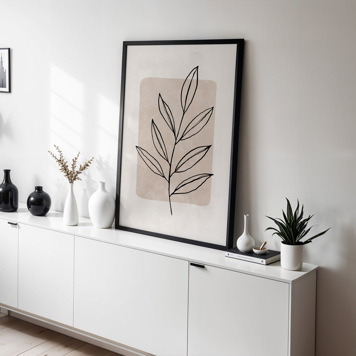 Black line slender leaf on beige panel print in a black frame leaning on a white sideboard with black and white vases