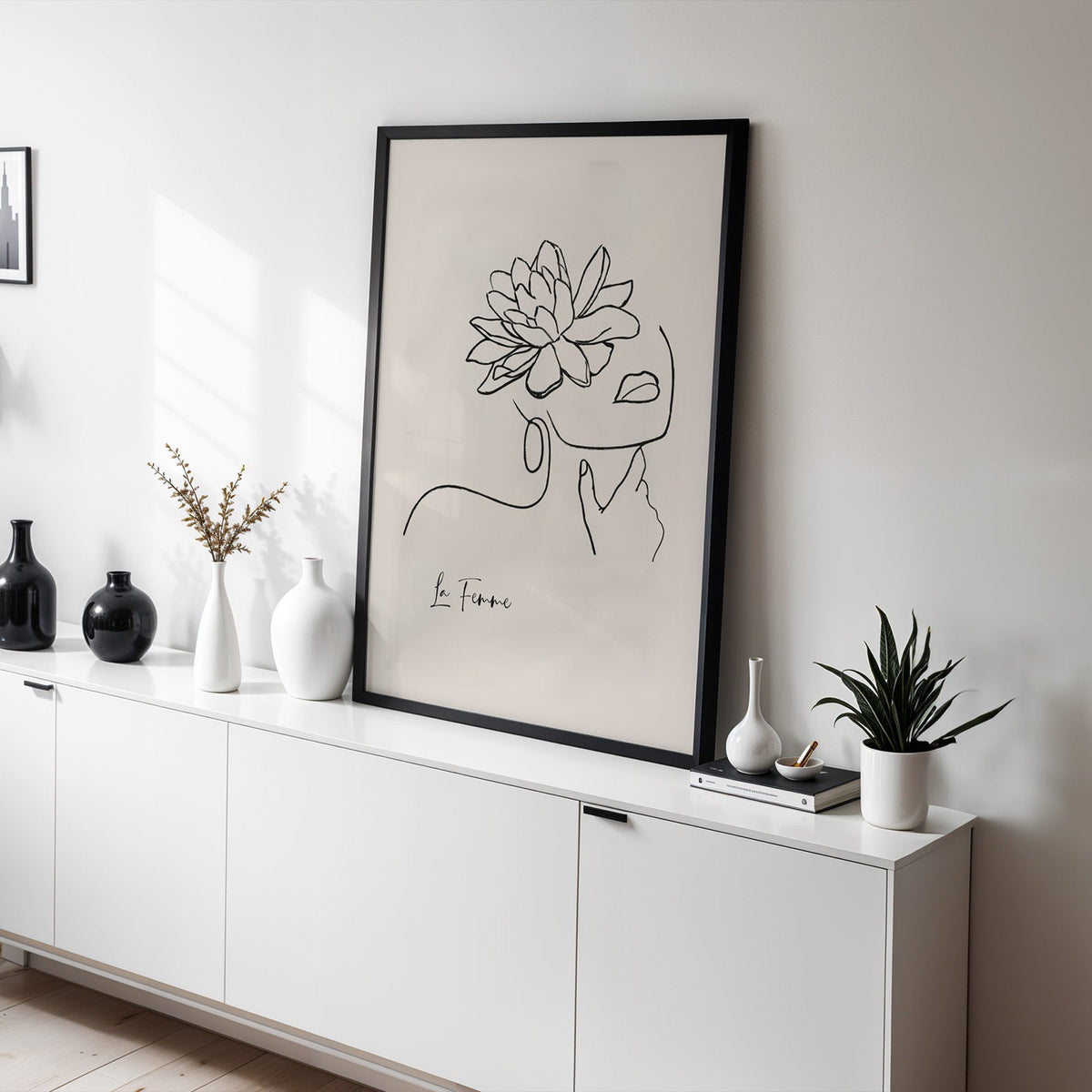 Woman face and flower black line art print in a black frame leaning on a white sideboard with black and white vases
