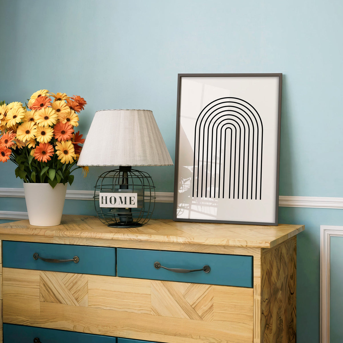 Black nested double line arches print in a charcoal frame on a teal dresser beside orange and yellow daisies