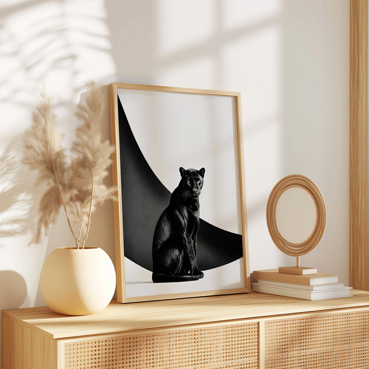 Seated black panther in front of black arc print in an oak frame on a rattan sideboard beside pampas grass