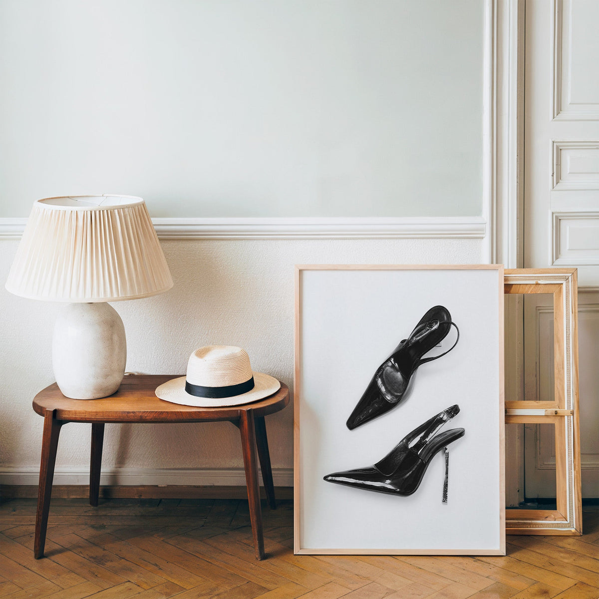 Black patent slingbacks with silver heels print in a pine frame on a wood floor beside a lamp and straw hat