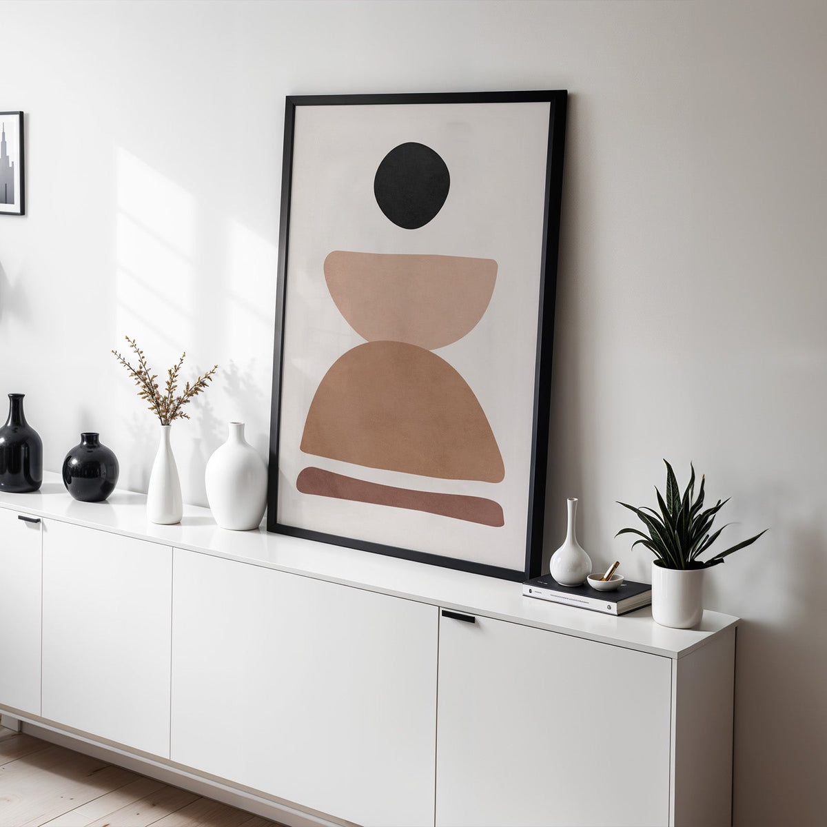 Black pebble and stacked stones mid century print in a black frame leaning on a white sideboard with black and white vases