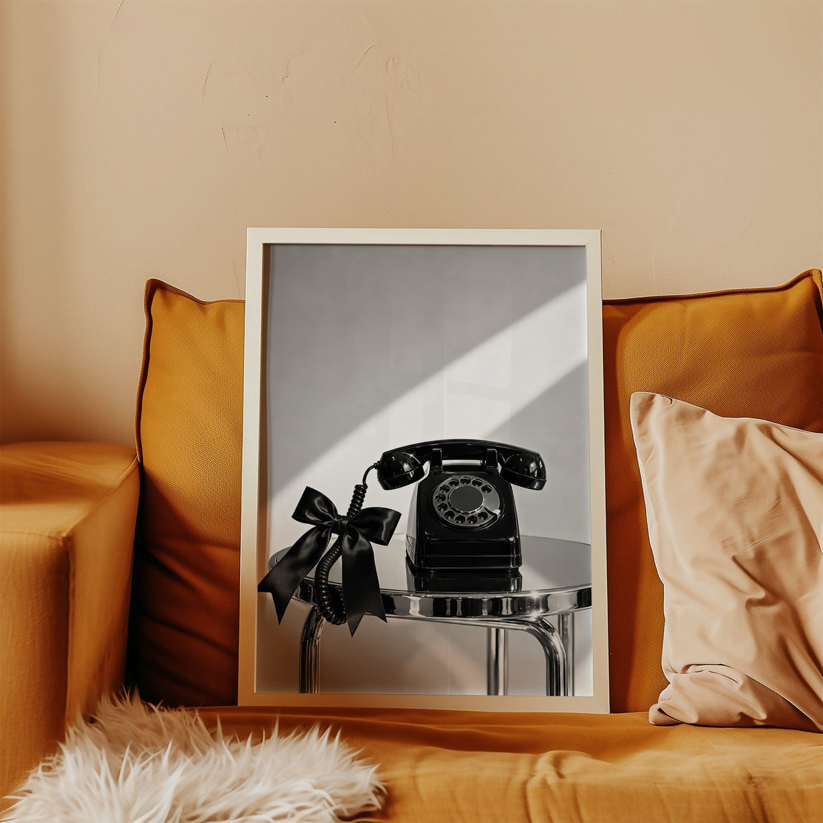 Black rotary phone with satin bow on chrome table print in a white frame leaning on a mustard velvet sofa