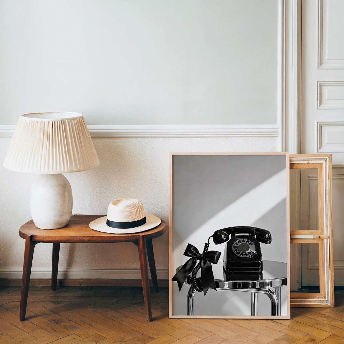 Black rotary phone with satin bow on chrome table print in a pine frame on a wood floor beside a lamp and straw hat