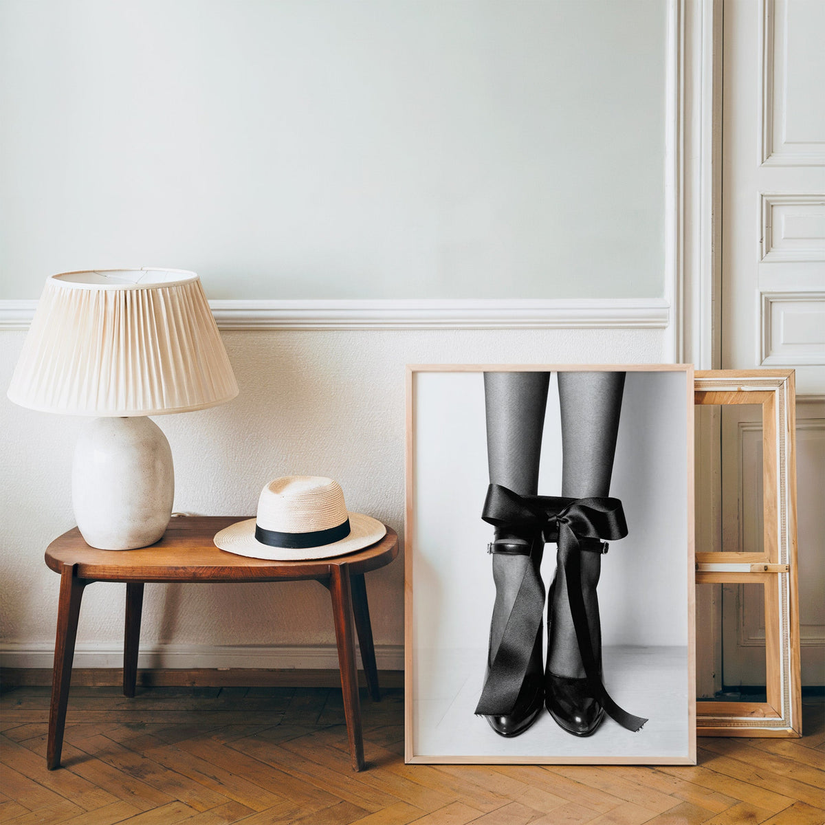 Heels with black satin bows tied at the ankle print in a pine frame on a wood floor beside a lamp and straw hat