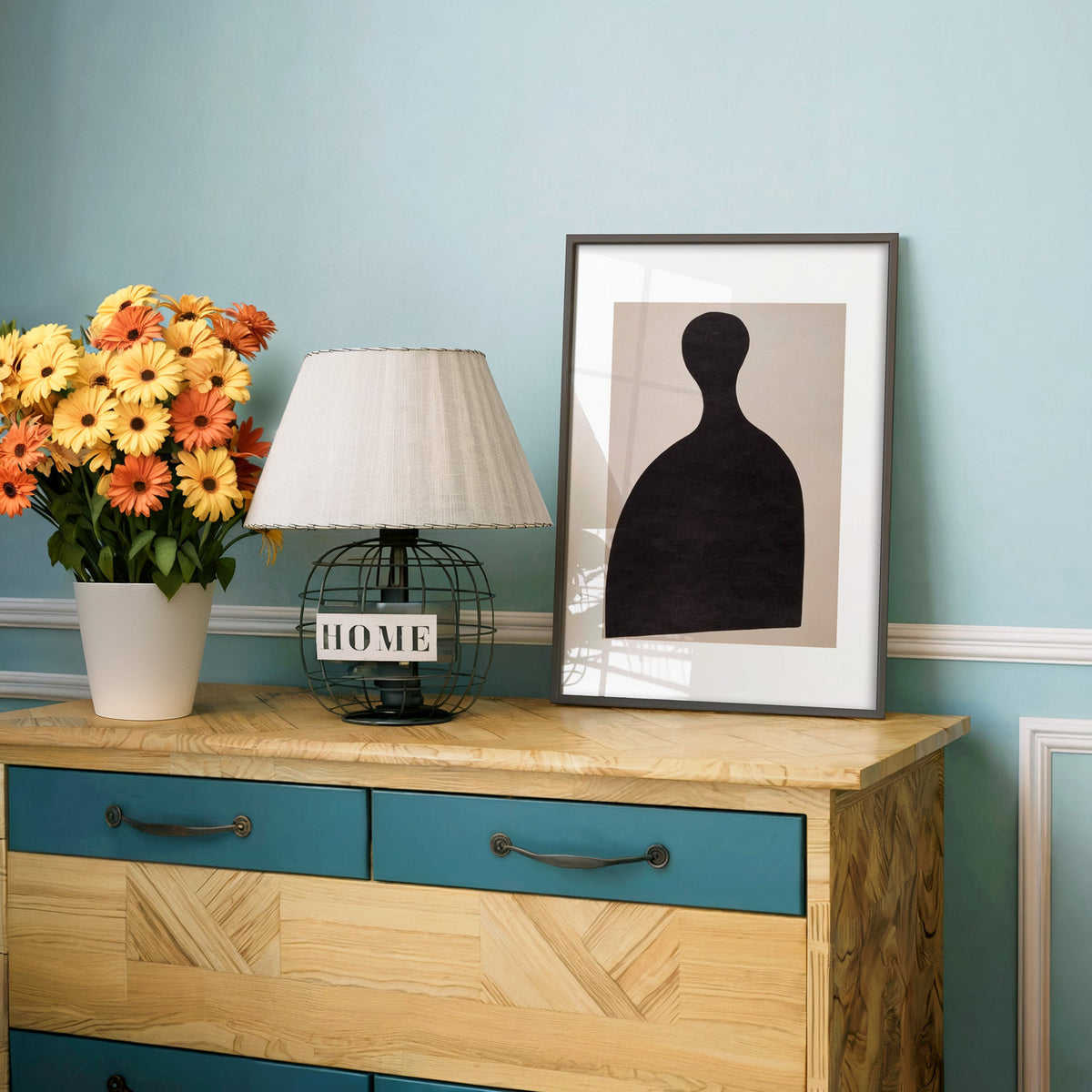 Black silhouette bust on blush panel print in a charcoal frame on a teal dresser beside orange and yellow daisies