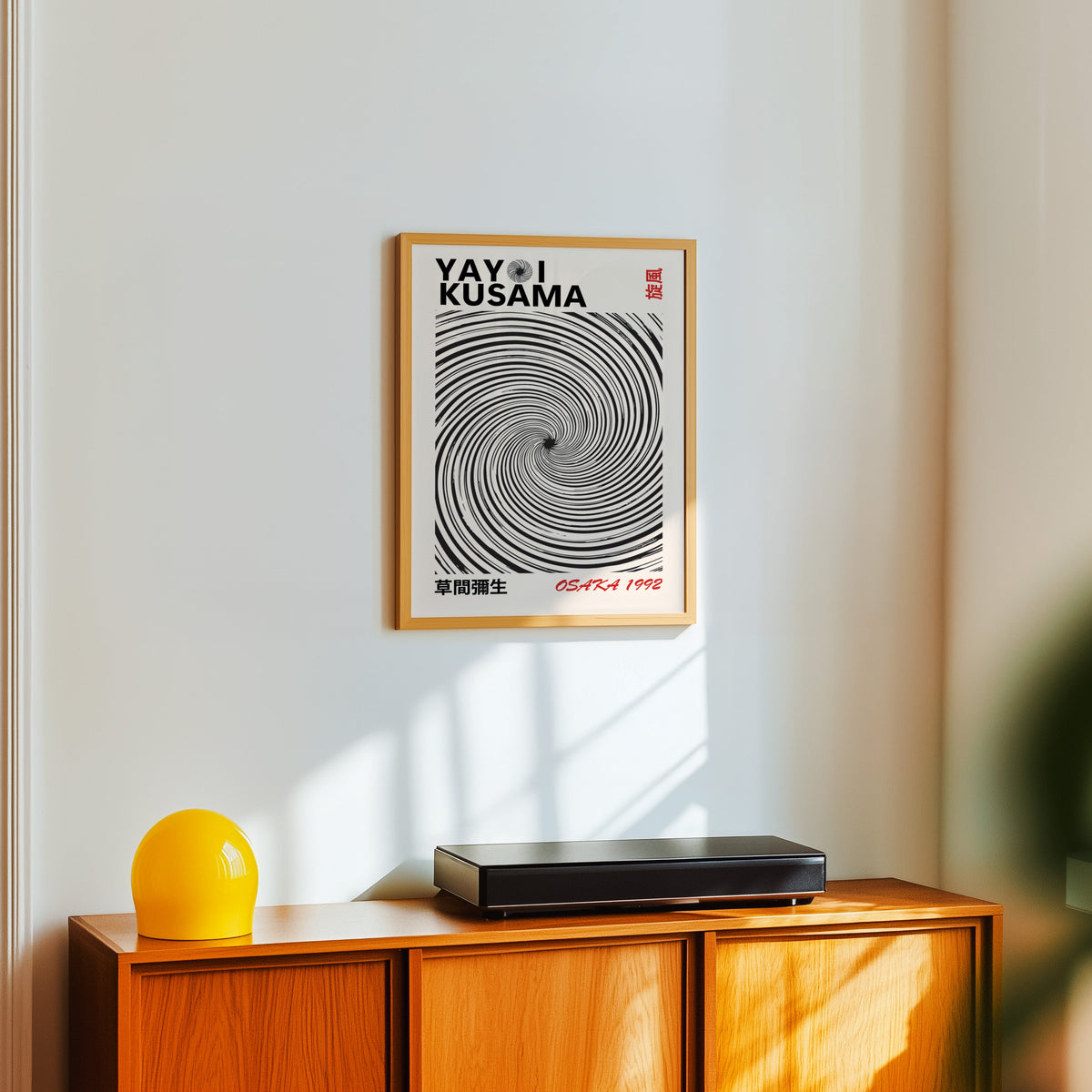Yayoi Kusama inspired black spiral whirlwind print in an oak frame over a teak sideboard with a yellow lamp