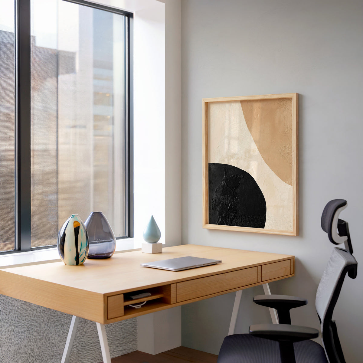 Black and tan textured shapes abstract print in a pale wood frame above a home office desk by a tall window