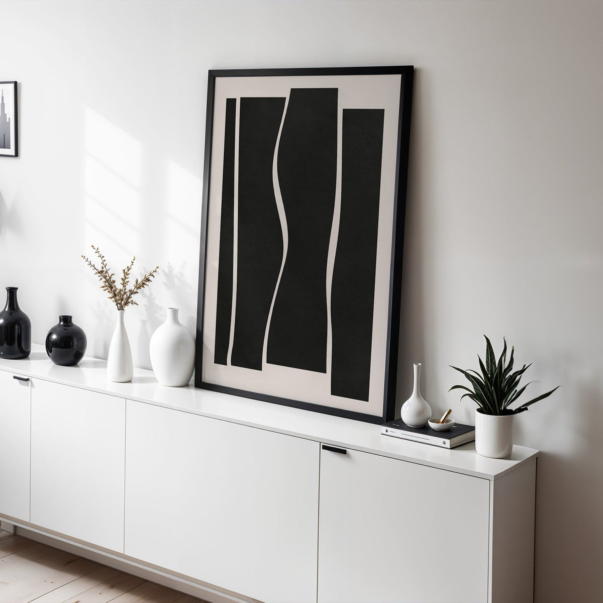 Black wavy vertical panels minimalist print in a black frame leaning on a white sideboard with black and white vases