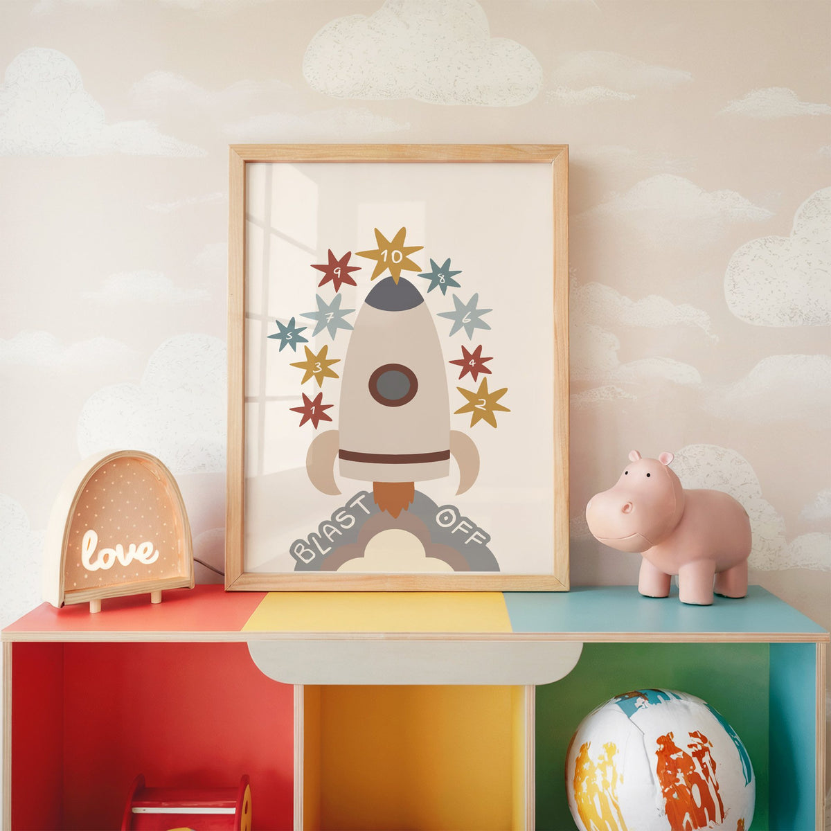 Blast off rocket countdown nursery print in an oak frame on a rainbow toy shelf against cloud wallpaper