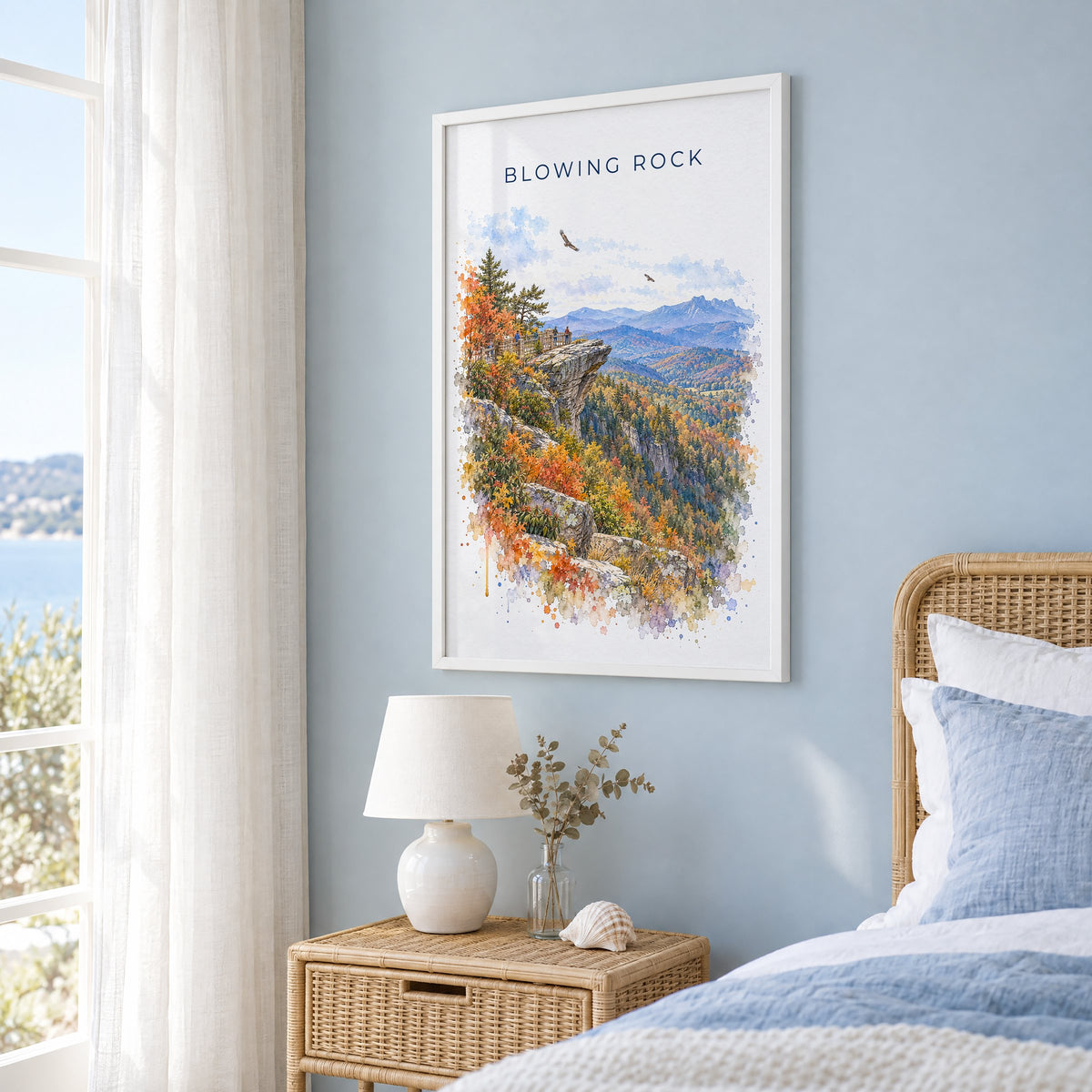 Blowing Rock watercolor print in a white frame on a pale blue wall beside a rattan bed in a coastal bedroom