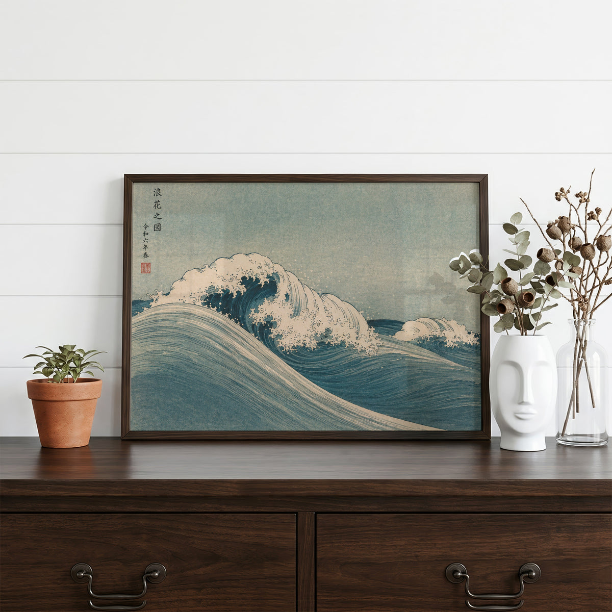 Blue crashing wave woodblock print in a dark walnut frame on a dark wood dresser beside a white face vase