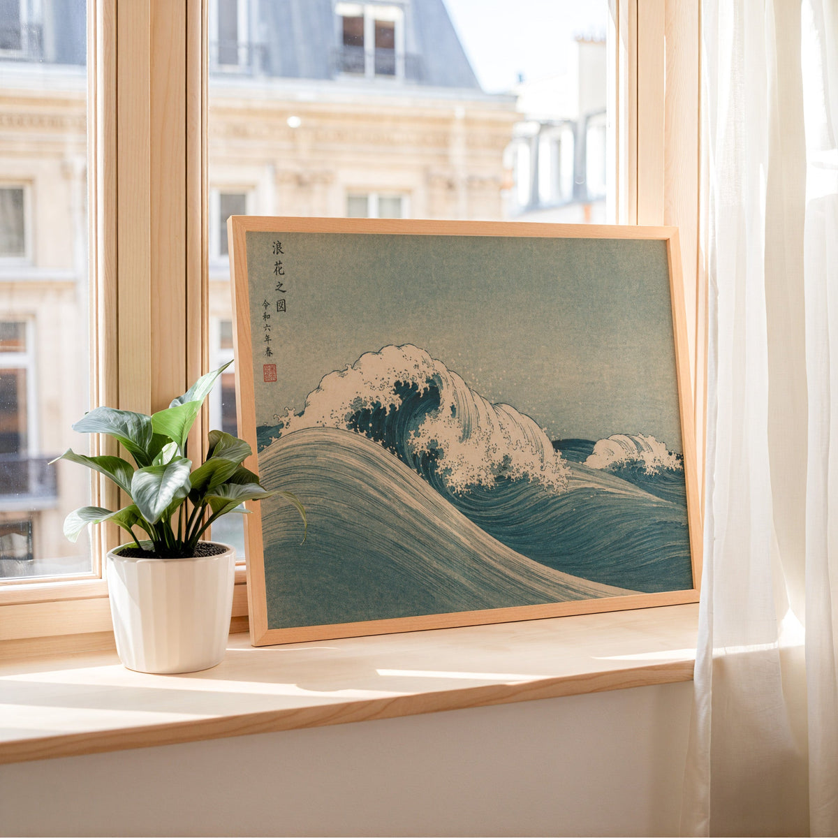 Blue crashing wave woodblock print in a light wood frame leaning on a sunny windowsill by sheer curtains