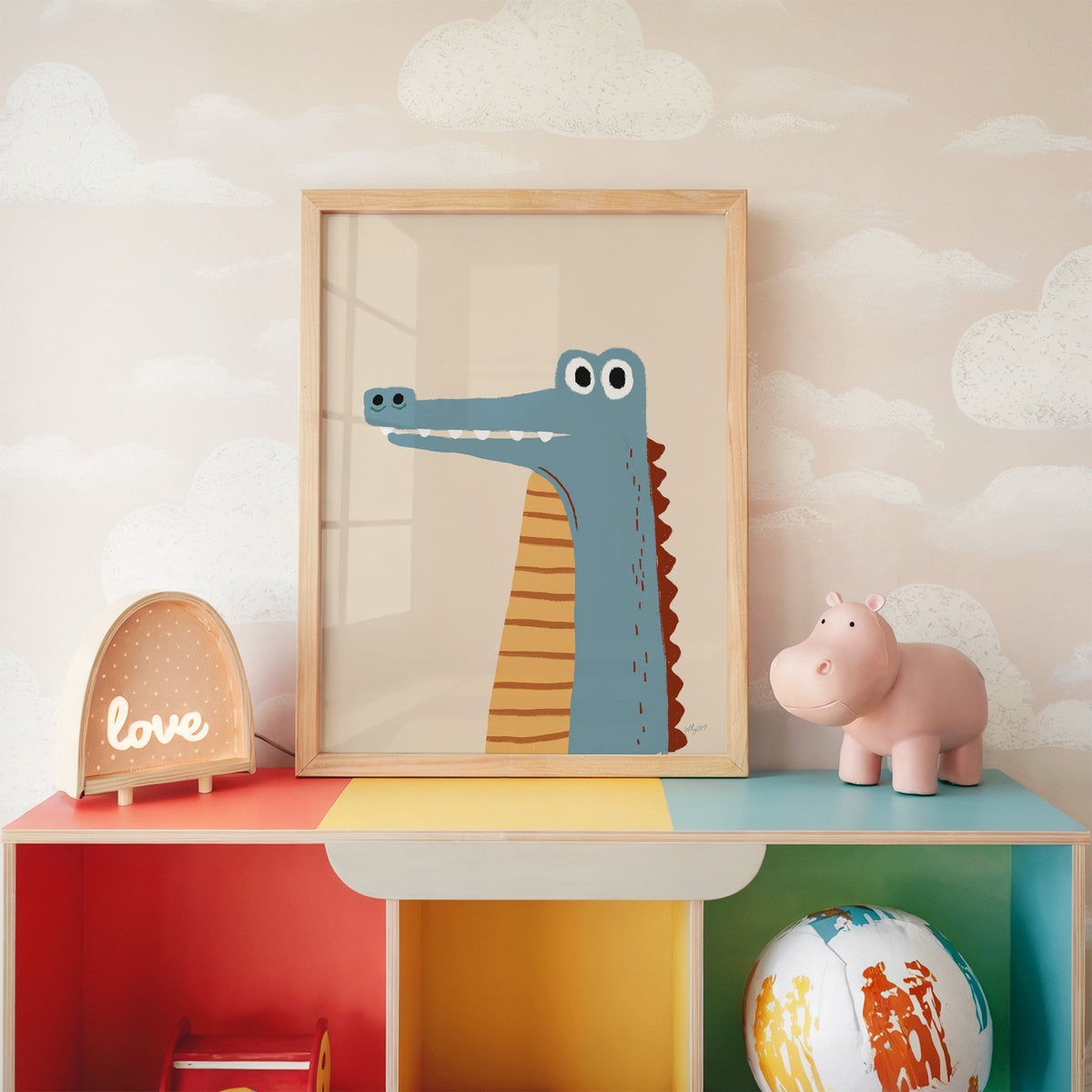 Upright blue crocodile on cream nursery print in an oak frame on a rainbow toy shelf against cloud wallpaper