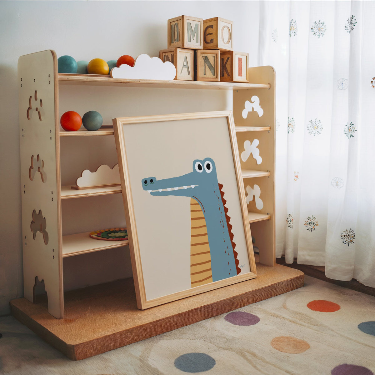 Upright blue crocodile on cream nursery print in an oak frame leaning on a toy shelf with wooden letter blocks