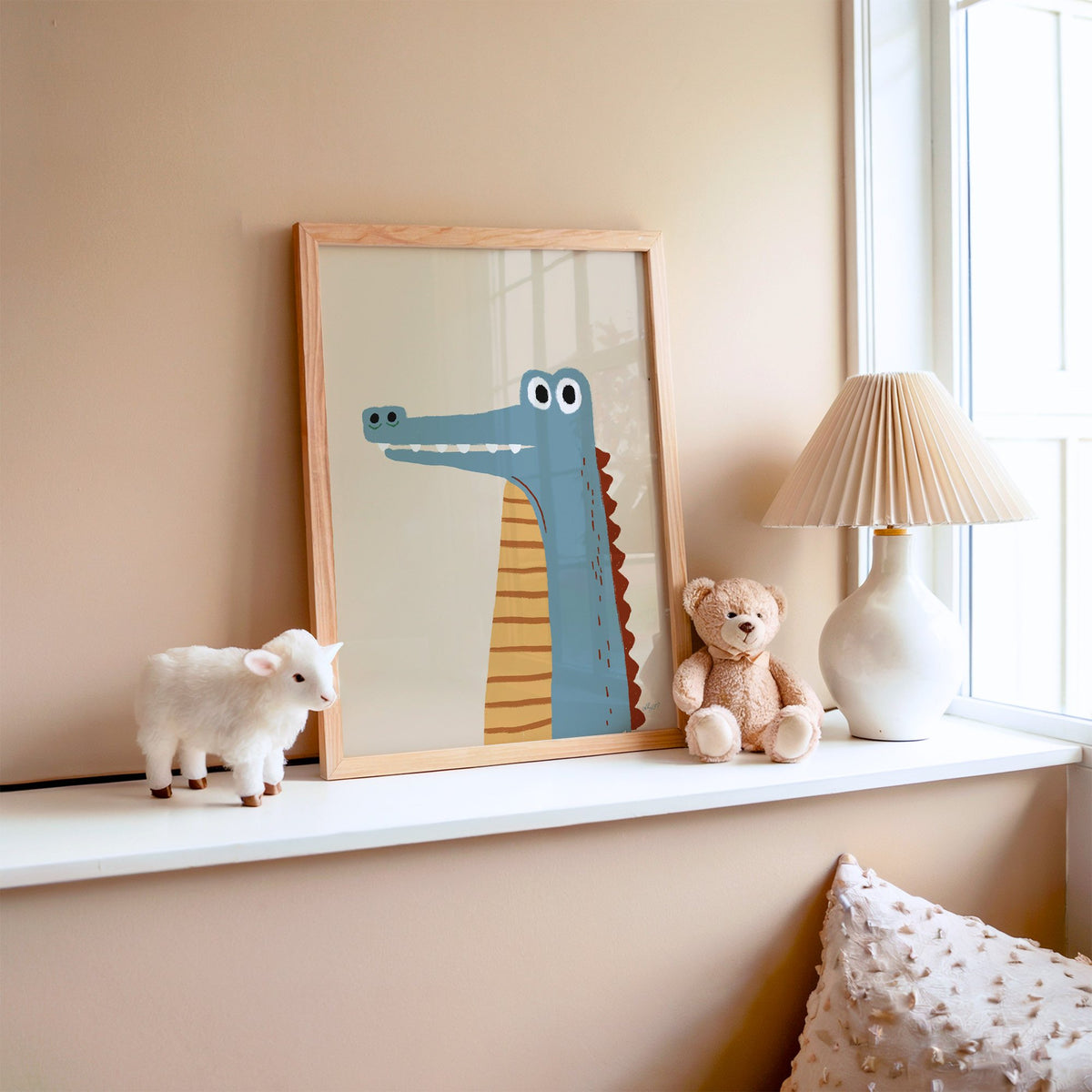 Upright blue crocodile on cream nursery print in an oak frame on a window ledge with a toy lamb and teddy