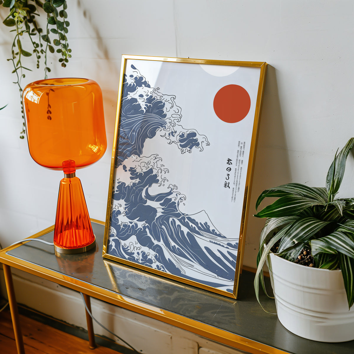 Blue Great Wave with red sun print shown in a gold frame on a console beside an orange glass lamp