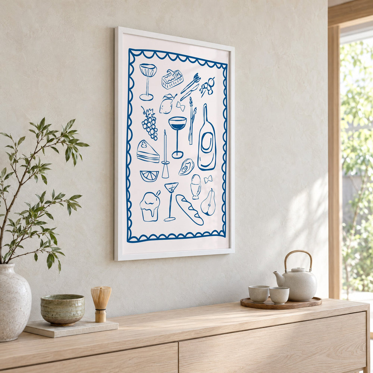 Blue line dinner party scalloped print in a white frame above a pale wood console with a matcha tea set