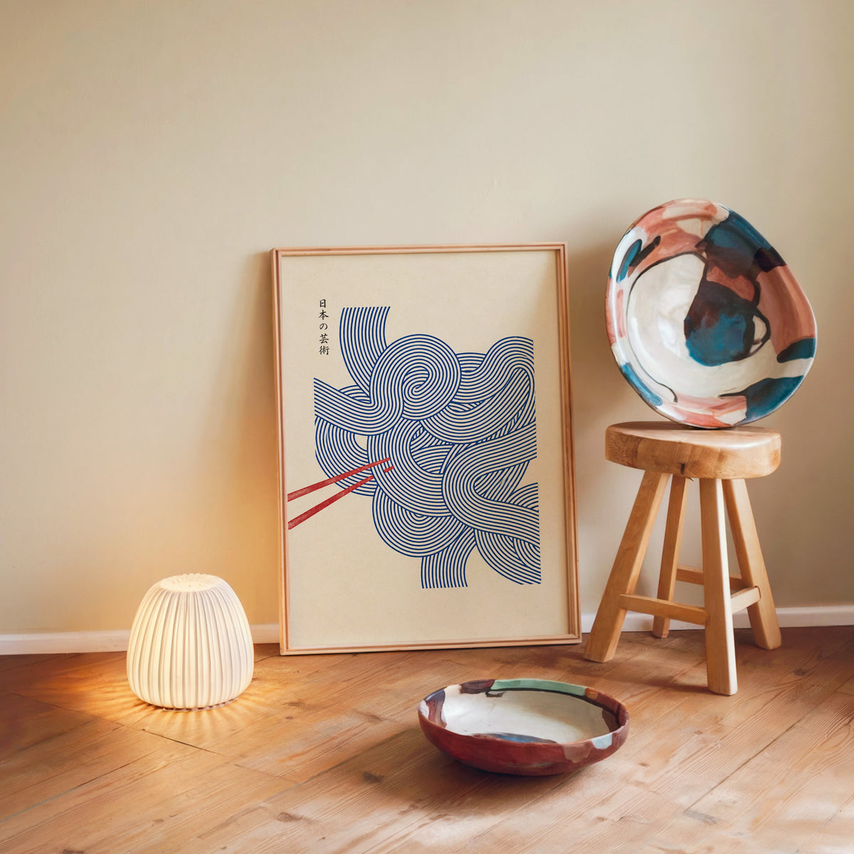 Blue line noodles poster in a light wood frame leaning on a wooden floor by a glowing lamp and stool
