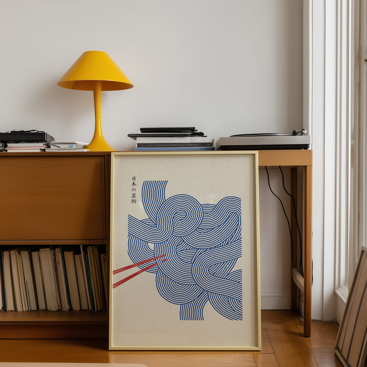 Blue line noodles poster shown in a gold frame leaning under a teak desk with a yellow lamp