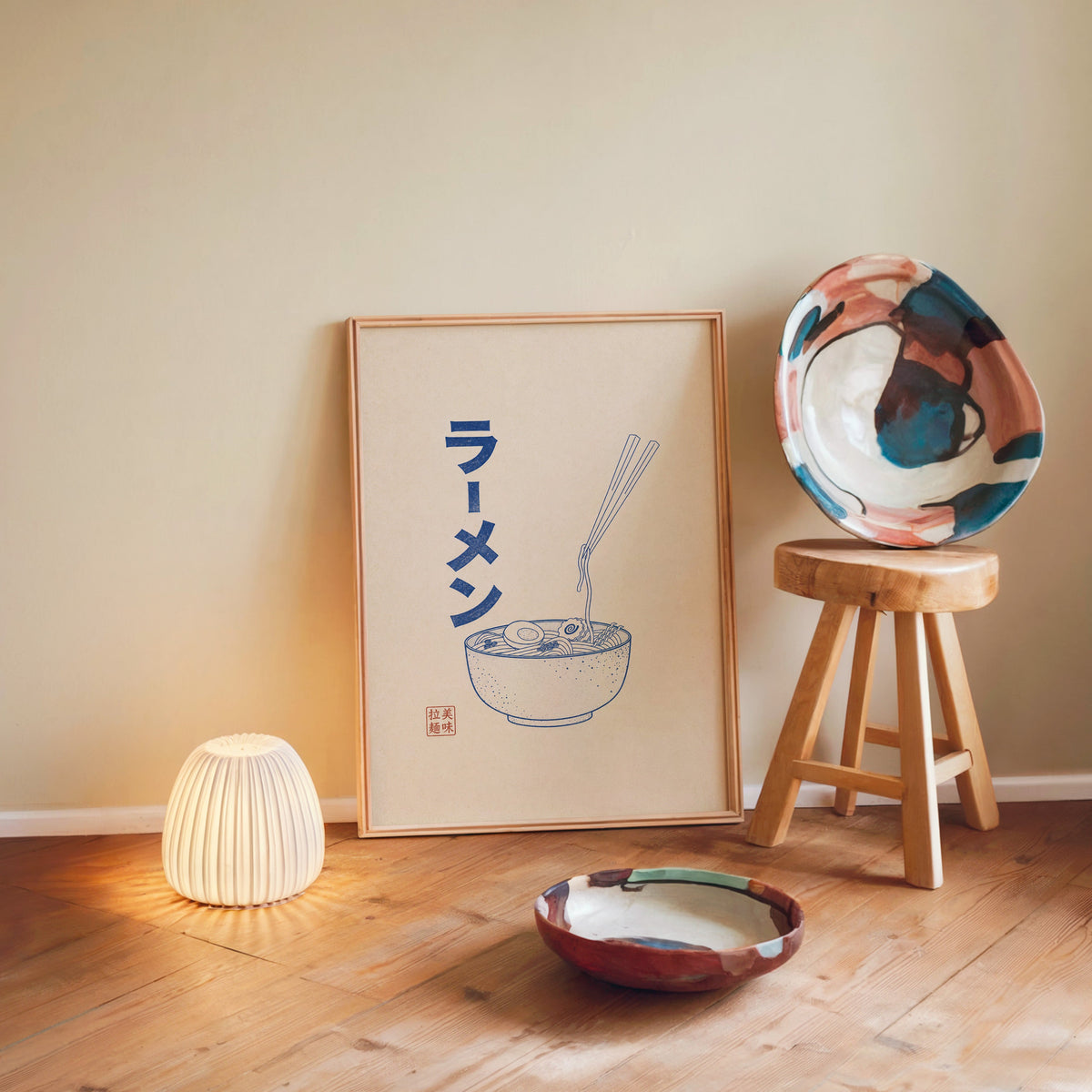 Blue line ramen bowl poster in a light wood frame leaning on a wooden floor by a glowing lamp and stool