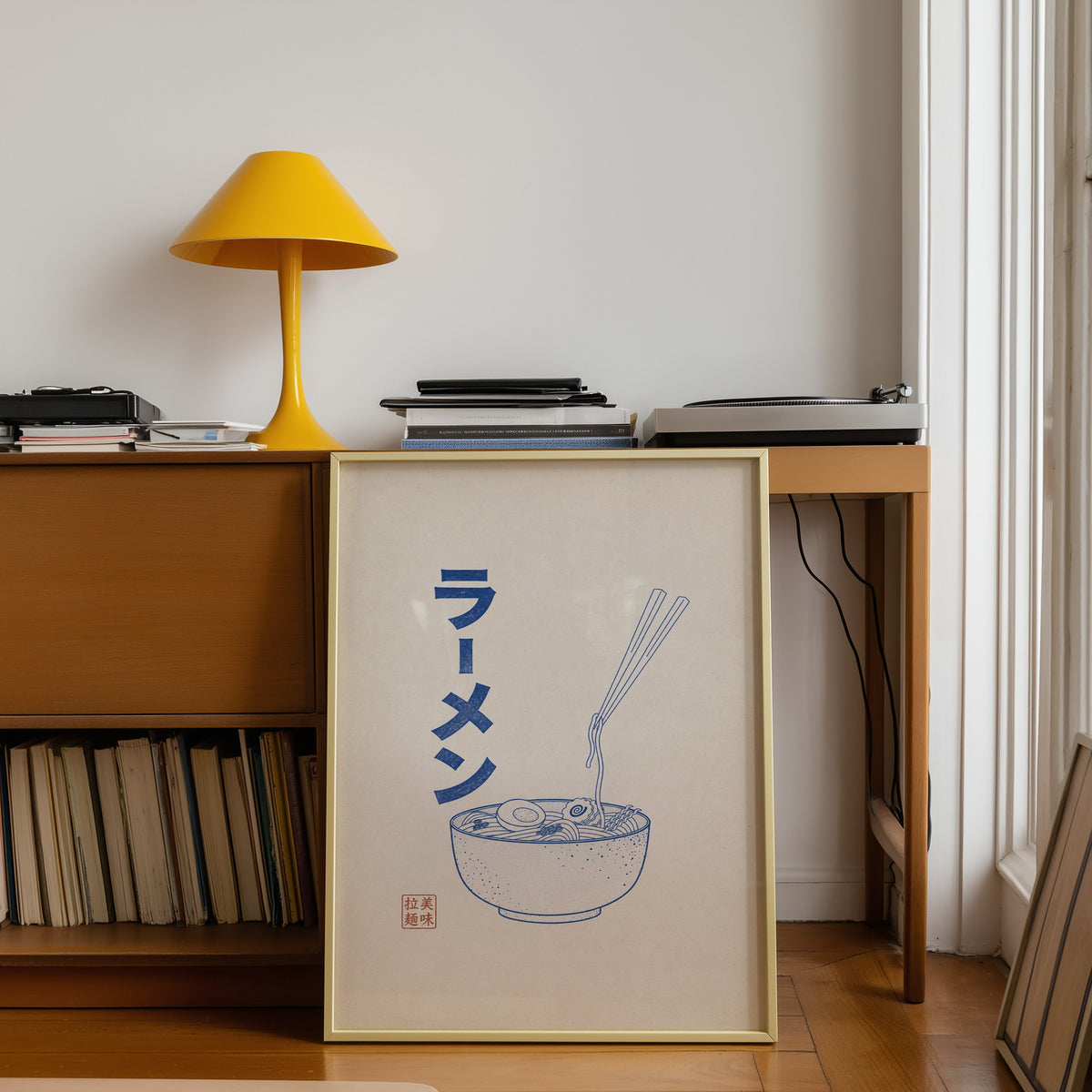 Blue line ramen bowl poster shown in a gold frame leaning under a teak desk with a yellow lamp