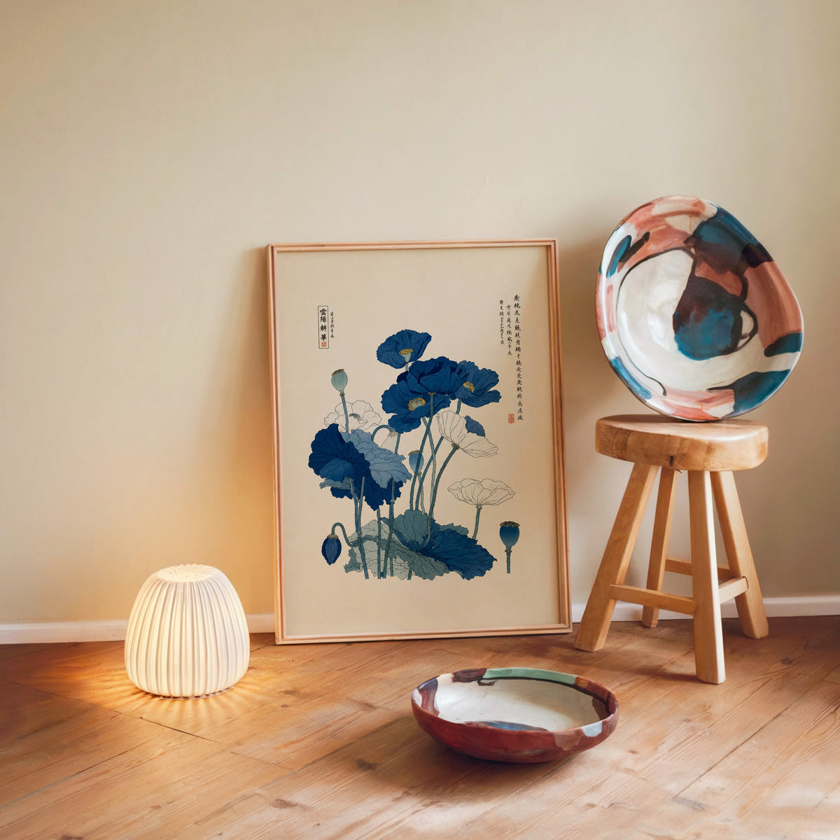 Blue and white poppies poster in a light wood frame leaning on a wooden floor by a glowing lamp and stool