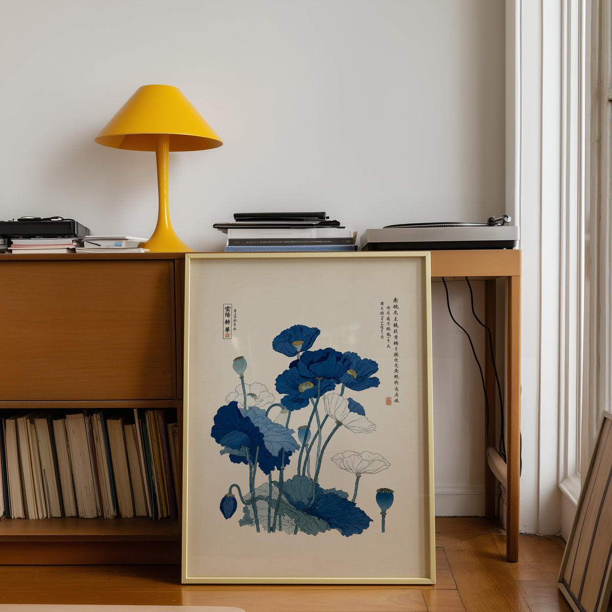 Blue and white poppies poster shown in a gold frame leaning under a teak desk with a yellow lamp